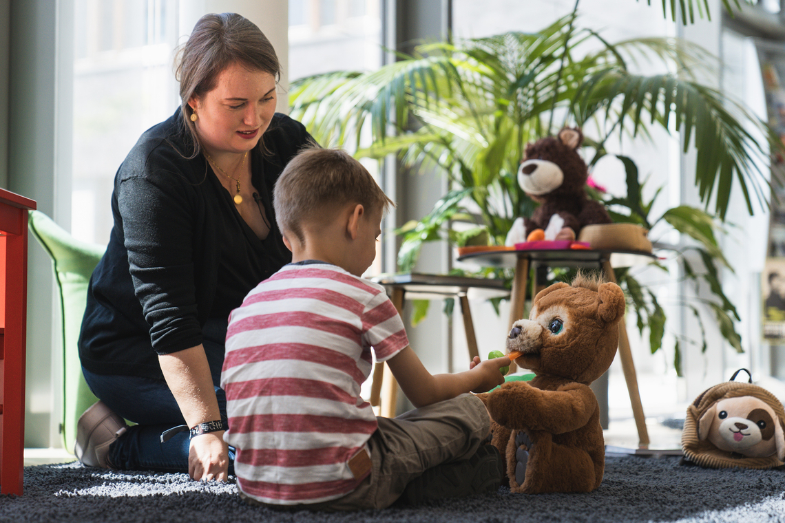 Eine Frau sitzt mit Kita-Kind auf dem Boden und spielt mit Kuscheltieren.